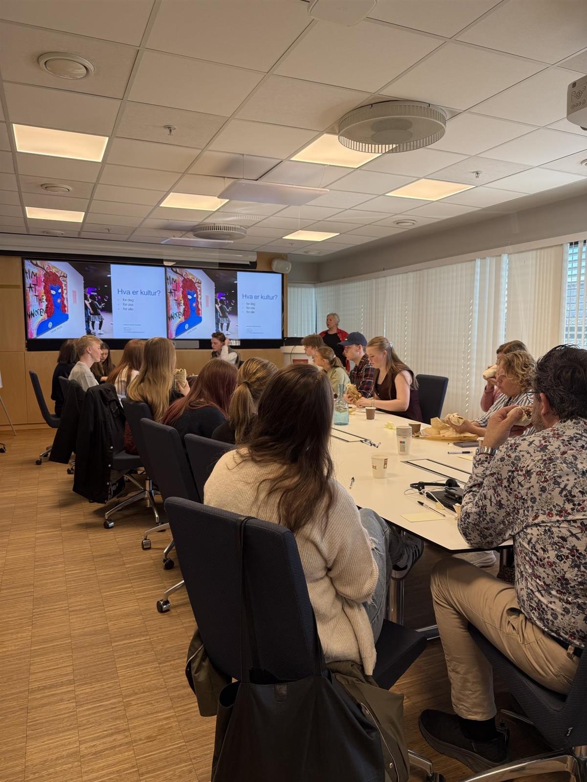 Representanter fra alle rådene som er samlet rundt bordet i formannskapssalen, i enden av rommet foran to skjermer står en rådgiver å forteller om kultur. - Klikk for stort bilde